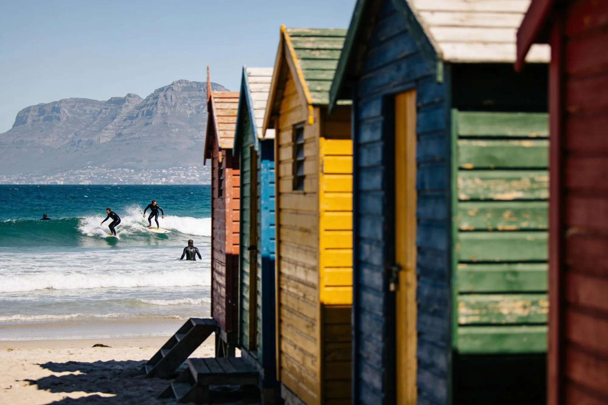 La hilera de casetas de baño victorianas de colores en la playa de Muizenberg con surfistas en el agua
